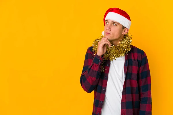 头戴圣诞礼帽的年轻的高加索人 带着一份黄色背景的礼物 面面相觑 脸上带着怀疑和怀疑的表情 — 图库照片