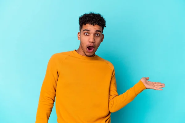 Jovem Afro Americano Encaracolado Homem Isolado Azul Impressionado Segurando Espaço — Fotografia de Stock