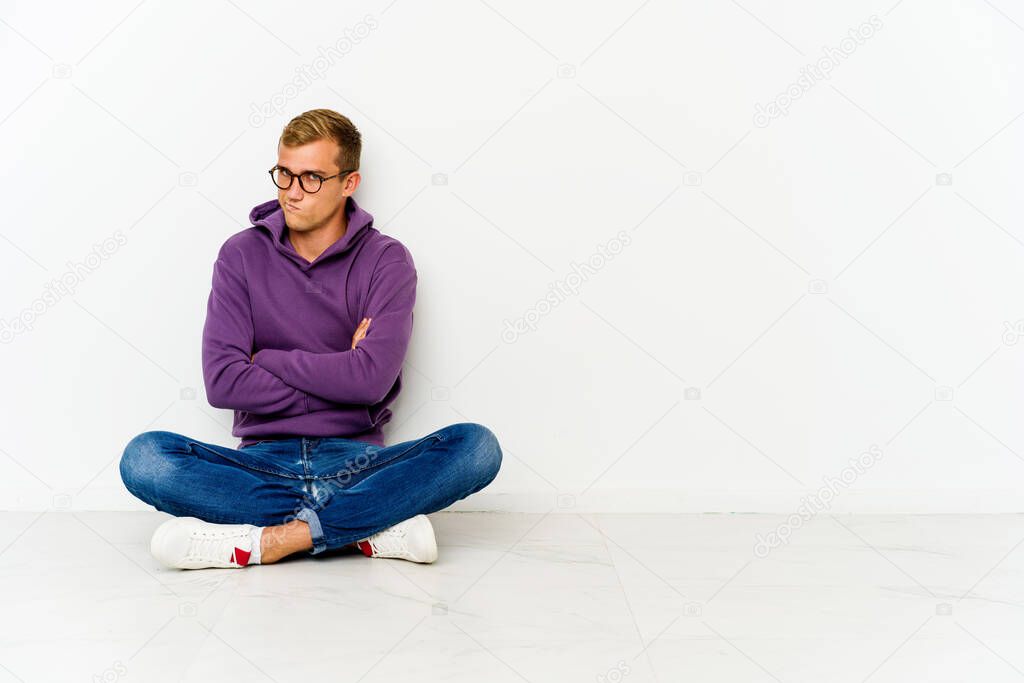 Young caucasian man sitting on the floor frowning face in displeasure, keeps arms folded.