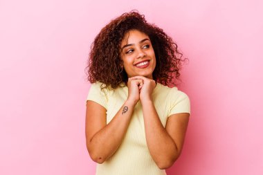 Pembe arka planda izole edilmiş genç Afrikalı Amerikalı kadın ellerini çenesinin altında tutuyor, mutlu bir şekilde yan tarafa bakıyor..