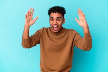 Genç Afro-Amerikalı kıvırcık adam mavi üzerine izole edilmiş. Hoş bir sürpriz alıyor, heyecanlı ve el kaldırıyor..