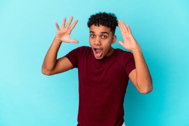 Genç Afro-Amerikalı kıvırcık adam mavi üzerine izole edilmiş zaferi ya da başarıyı kutluyor, şaşırıyor ve şok oluyor..