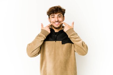 Beyaz arka plandaki genç Arap adam gülümsüyor, parmaklarını ağıza doğrultuyor..