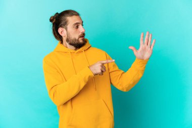 Uzun saçlı genç adam gülümsüyor. Beş numarayı parmaklarıyla gösteriyor..