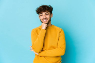 Mavi arka plandaki genç Arap adam gülümsüyor mutlu ve kendinden emin, elleriyle çenesine dokunuyor..