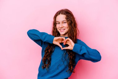 Pembe arka planda izole edilmiş küçük beyaz kız gülümsüyor ve elleriyle kalp şeklini gösteriyor..