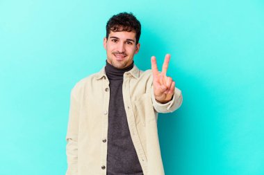 Mavi arka planda izole edilmiş genç beyaz adam zafer işareti gösteriyor ve gülümsüyor..