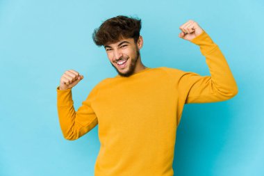 Mavi arka planda genç bir Arap özel bir günü kutluyor, atlıyor ve enerji ile kollarını kaldırıyor..