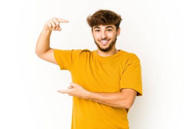 Beyaz arka plandaki genç Arap adam işaret parmaklarıyla küçük bir şey tutuyor, gülümsüyor ve kendinden emin..