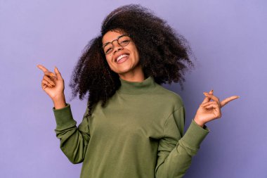 Genç Afro-Amerikalı kıvırcık kadın, mor arka planda izole edilmiş, farklı fotokopi alanlarına bakıyor, birini seçiyor, parmaklarıyla gösteriyor..