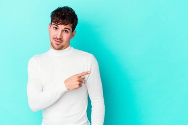 Mavi arka planda izole edilmiş genç beyaz adam gülümsüyor ve boş alanda bir şeyler gösteriyor..