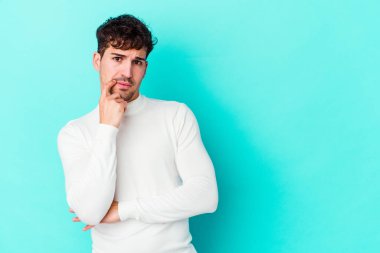 Mavi arka planda izole edilmiş genç beyaz adam şüpheli ve şüpheci bir ifadeyle yana bakıyor..