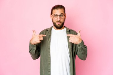 Uzun saçlı genç adam parmaklarıyla işaret ederek şaşırmış görünüyor, geniş bir gülümsemeyle..