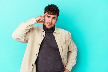 Mavi arka planda izole edilmiş genç beyaz bir adam şakağı parmağıyla işaret ediyor, düşünüyor ve bir göreve odaklanıyor..