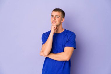 Genç beyaz yakışıklı adam gülümsüyor mutlu ve kendinden emin, çenesine elle dokunuyor..