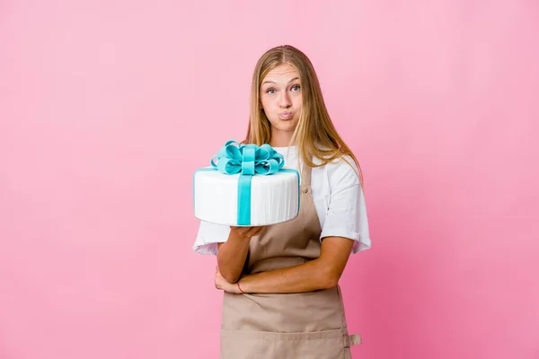 Young russian baker woman holding a delicious cake blows cheeks, has tired expression. Facial expression concept.
