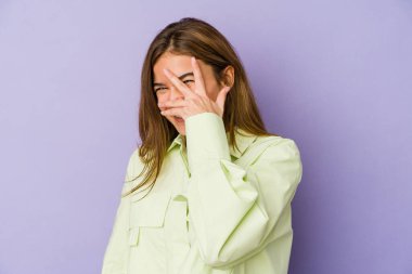 Mor arka planda genç, sıska, beyaz bir kız kameraya parmaklarıyla göz kırpıyor, utanarak yüzünü kapatıyor..