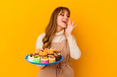 Sarı arka planda izole edilmiş genç pasta şefi kadın bağırıyor ve avuç içini açık tutuyor..