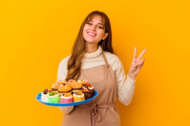 Sarı arka planda izole edilmiş genç pasta şefi kadın iki numarayı parmaklarıyla gösteriyor..