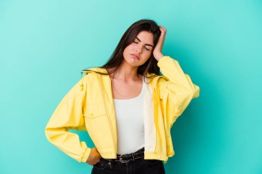 Mavi arka planda izole edilmiş genç beyaz kadın yorgun ve uykulu ellerini başının üstünde tutuyor..