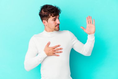 Kafkasyalı genç adam mavi arka planda izole edilmiş yemin ediyor, elini göğsüne koyuyor..