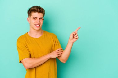 Mavi arka planda izole edilmiş genç beyaz adam işaret parmağıyla işaret ederek gülümsüyor..