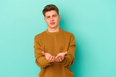 Kafkasyalı genç adam mavi arka planda izole edilmiş. Elinde avuç içi olan bir şey var ve kameraya sunuyor..