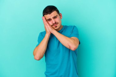 Mavi arka planda izole edilmiş genç beyaz adam esniyor. Ağzı eliyle kapatan yorgun bir hareket sergiliyor..