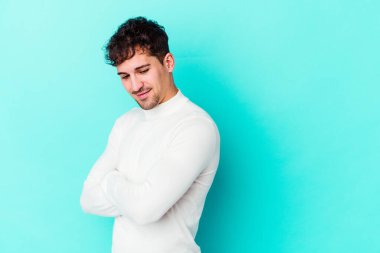 Mavi arka planda izole edilmiş genç beyaz adam kollarını kavuşturarak gülümsüyor..