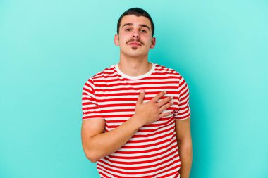 Kafkasyalı genç adam mavi arka planda izole edilmiş yemin ediyor, elini göğsüne koyuyor..