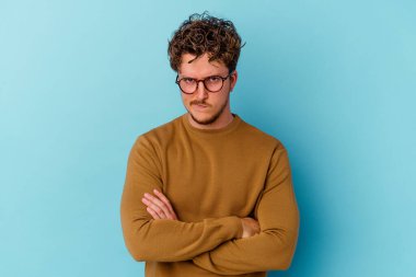 Gözlük takan, mavi arka planda soyutlanmış, hoşnutsuzca kaşlarını çatan, kollarını kavuşturan genç beyaz bir adam..