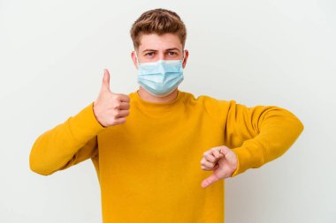 Koronavirüs maskesi takan genç adam beyaz arka planda izole edilmiş baş parmağını kaldırıyor ve baş parmağını aşağı gösteriyor, zor seçim konsepti