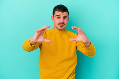 Kafkasyalı genç adam mavi arka planda izole edilmiş. Elinde avuç içi olan bir şey var ve kameraya sunuyor..