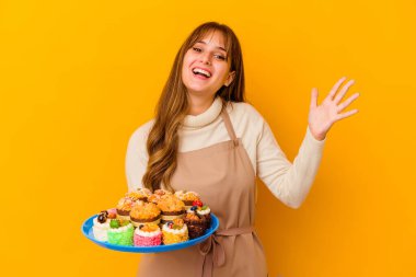 Sarı arka planda izole edilmiş genç pasta şefi kadın hoş bir sürpriz alıyor, heyecanlı ve el kaldırıyor..