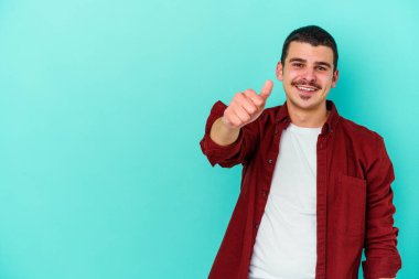 Mavi arka planda izole edilmiş genç beyaz adam gülümsüyor ve başparmağını kaldırıyor.