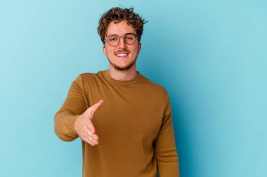 Mavi arka planda izole edilmiş gözlük takan genç beyaz adam selamlama jesti olarak kameraya elini uzatıyor..