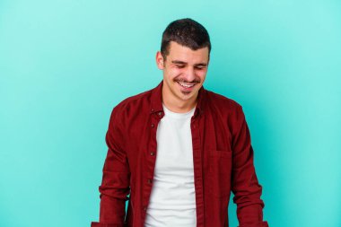 Mavi arka planda izole edilmiş genç beyaz adam gülüyor ve gözlerini kapıyor, rahatlamış ve mutlu hissediyor..