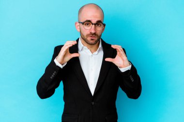 Genç beyaz, kel bir adam mavi arka planda izole edilmiş. Elinde avuç içi olan bir şey var. Kameraya teklif ediyor..