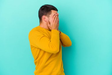 Mavi arka planda izole edilmiş genç beyaz adam elleriyle gözlerini kapatmaktan korkuyor..