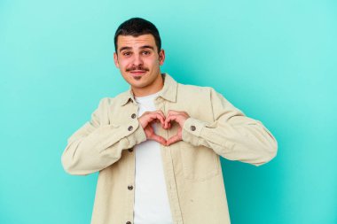 Mavi arka planda izole edilmiş genç beyaz adam gülümsüyor ve elleriyle kalp şeklini gösteriyor..