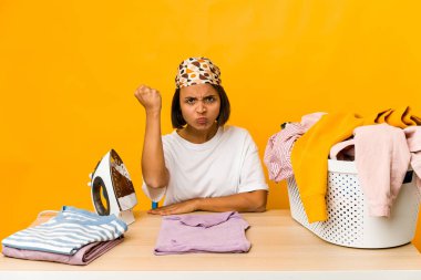 Giysileri ütüleyen genç İspanyol kadın kameraya yumruk gösteriyor, agresif yüz ifadesi var..