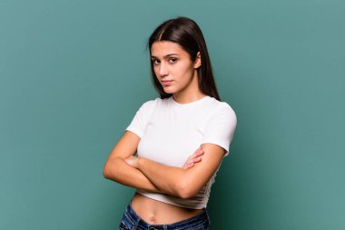 Mavi arka planda izole edilmiş genç Hintli kadın hoşnutsuzca kaşlarını çatmış, kollarını kavuşturmuş..