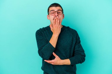 Mavi arka planda izole edilmiş genç beyaz adam esniyor. Ağzı eliyle kapatan yorgun bir hareket sergiliyor..