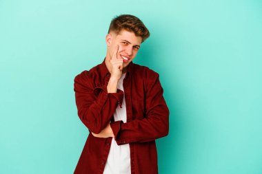 Mavi arka planda izole edilmiş genç beyaz adam gülümsüyor ve kendinden emin, elleriyle çenesine dokunuyor..