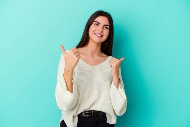 Kafkasyalı genç bir kadın mavi arka planda izole edilmiş. Baş parmağını kaldırıyor, gülümsüyor ve kendinden emin..