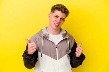 Sarı arka planda izole edilmiş genç beyaz adam iki parmağını da kaldırıyor, gülümsüyor ve kendinden emin..