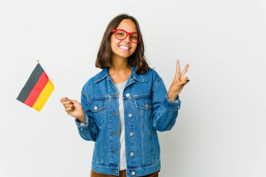 Beyaz arka planda izole edilmiş Alman bayrağı tutan genç Latin kadın zafer işareti gösteriyor ve gülümsüyor..