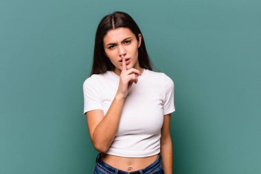 Genç Hintli kadın mavi arka planda izole edilmiş bir şekilde ya sır saklıyor ya da sessizlik istiyor..