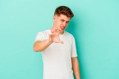 Mavi arka planda izole edilmiş genç beyaz adam pençelerini kedi taklidi yaparken gösteriyor, agresif bir hareket..