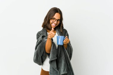 Beyaz arka planda battaniyeli genç Latin kadın gülümsüyor ve başparmağını kaldırıyor.